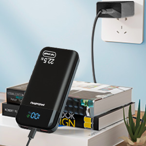 Cargador portátil negro enchufado a la toma de pared. La pantalla digital muestra el porcentaje de carga. Dispositivos electrónicos y planta en segundo plano.